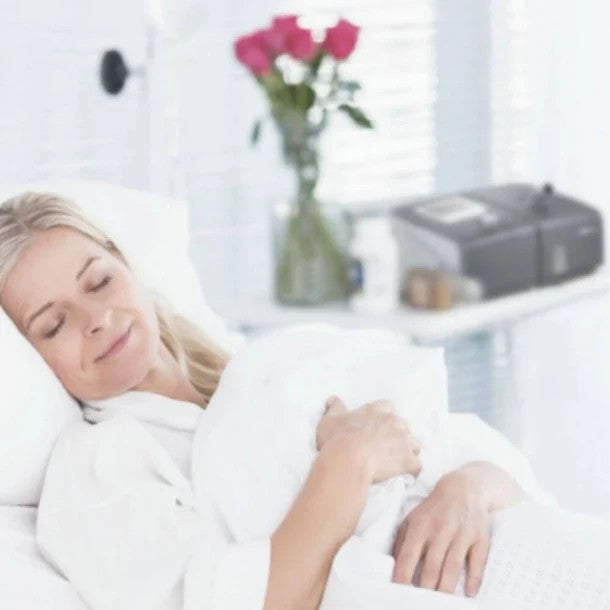 Woman lying down with a white blanket, smiling, in a bright room with flowers and a Resplus CPAP machine on her night stand